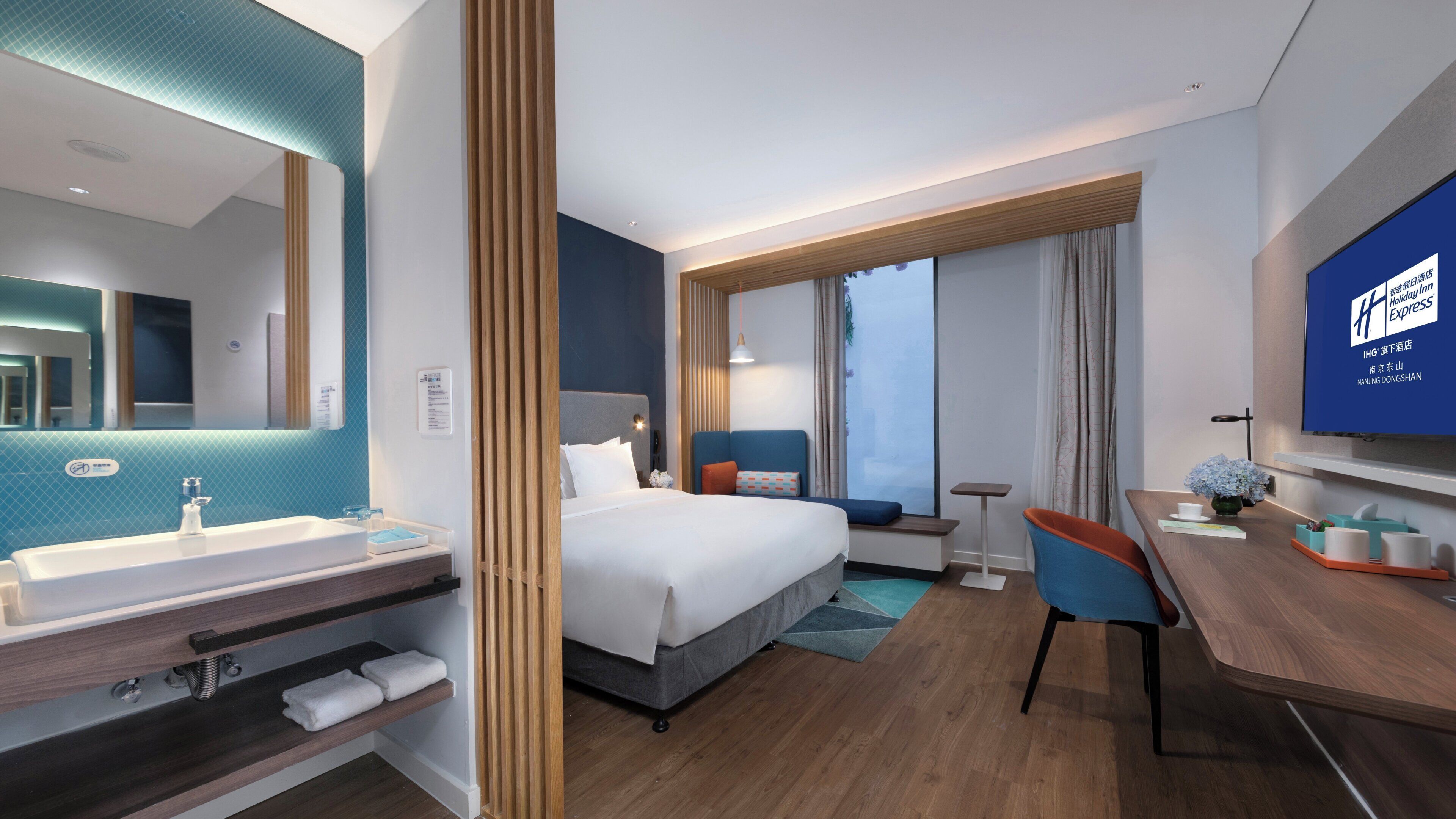 A hotel room with a bed, a desk, a chair, a TV, a bathroom with a sink and mirror, and a window with curtains.