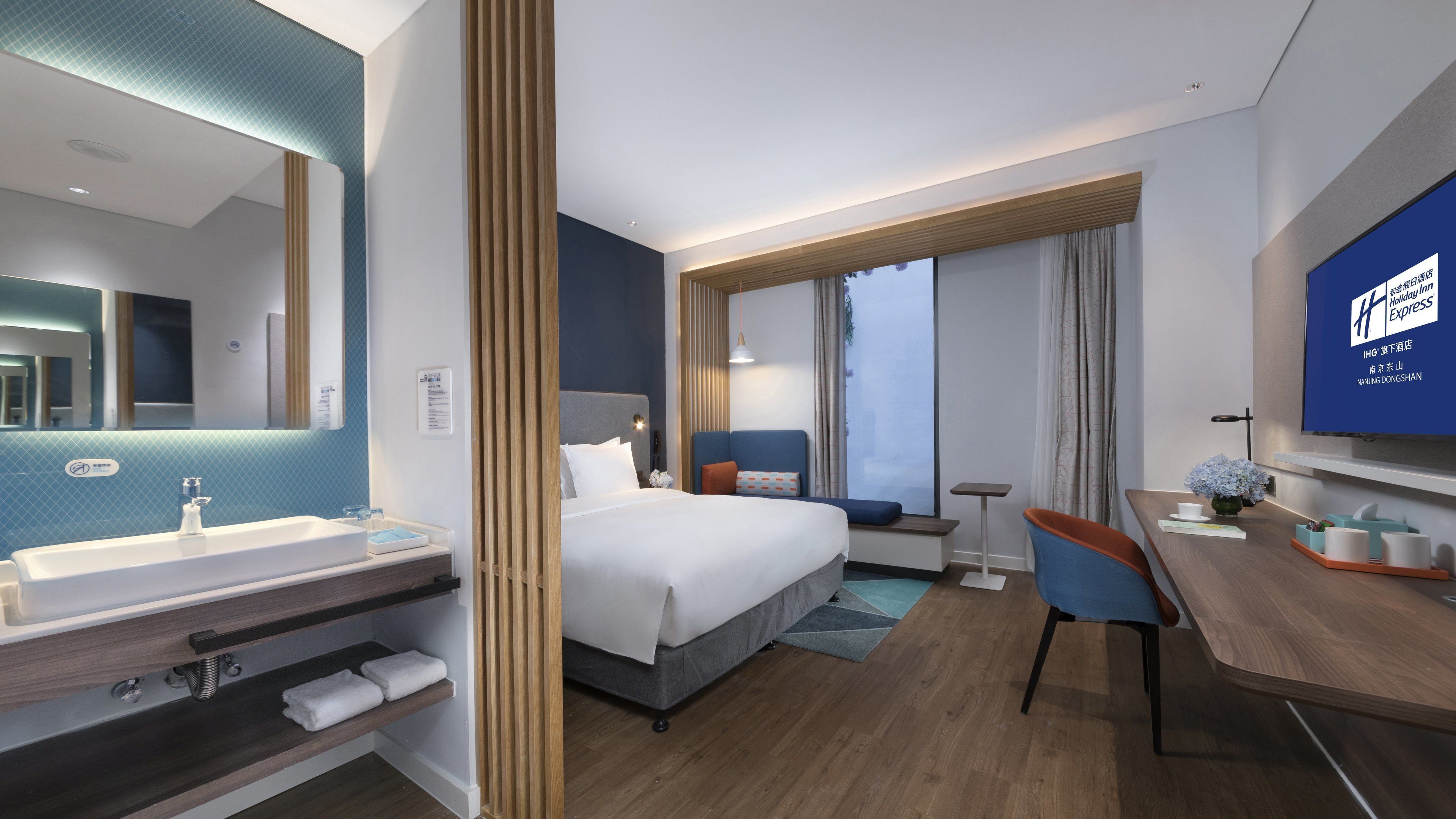 Une chambre d’hôtel comprenant un lit, un bureau, une chaise, une télévision, une salle de bain avec un lavabo et un miroir, et une fenêtre avec des rideaux.