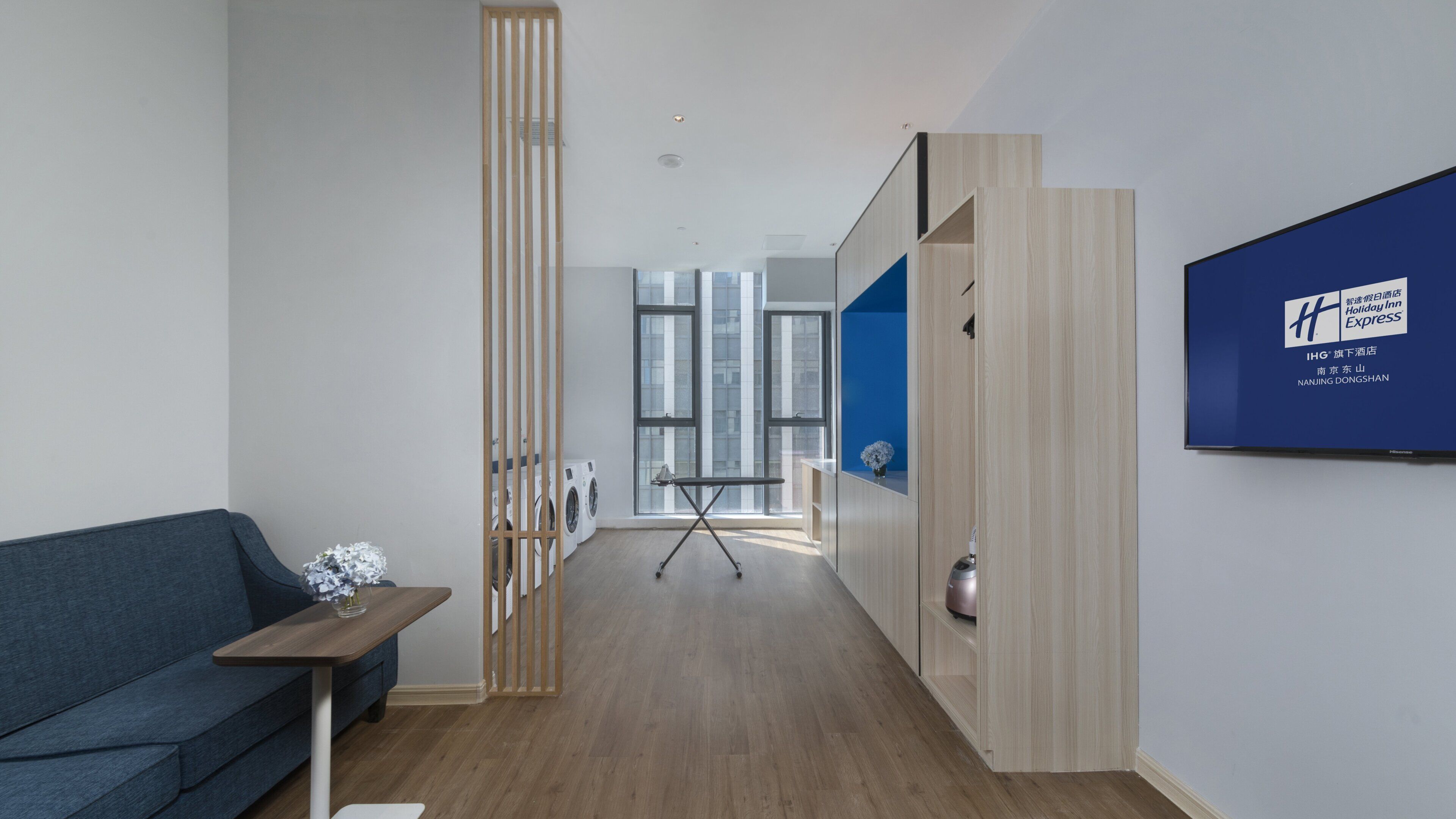 Habitación de hotel con un sofá azul, un separador de madera, un televisor de pantalla plana en la pared y vistas a la ciudad a través de amplios ventanales.