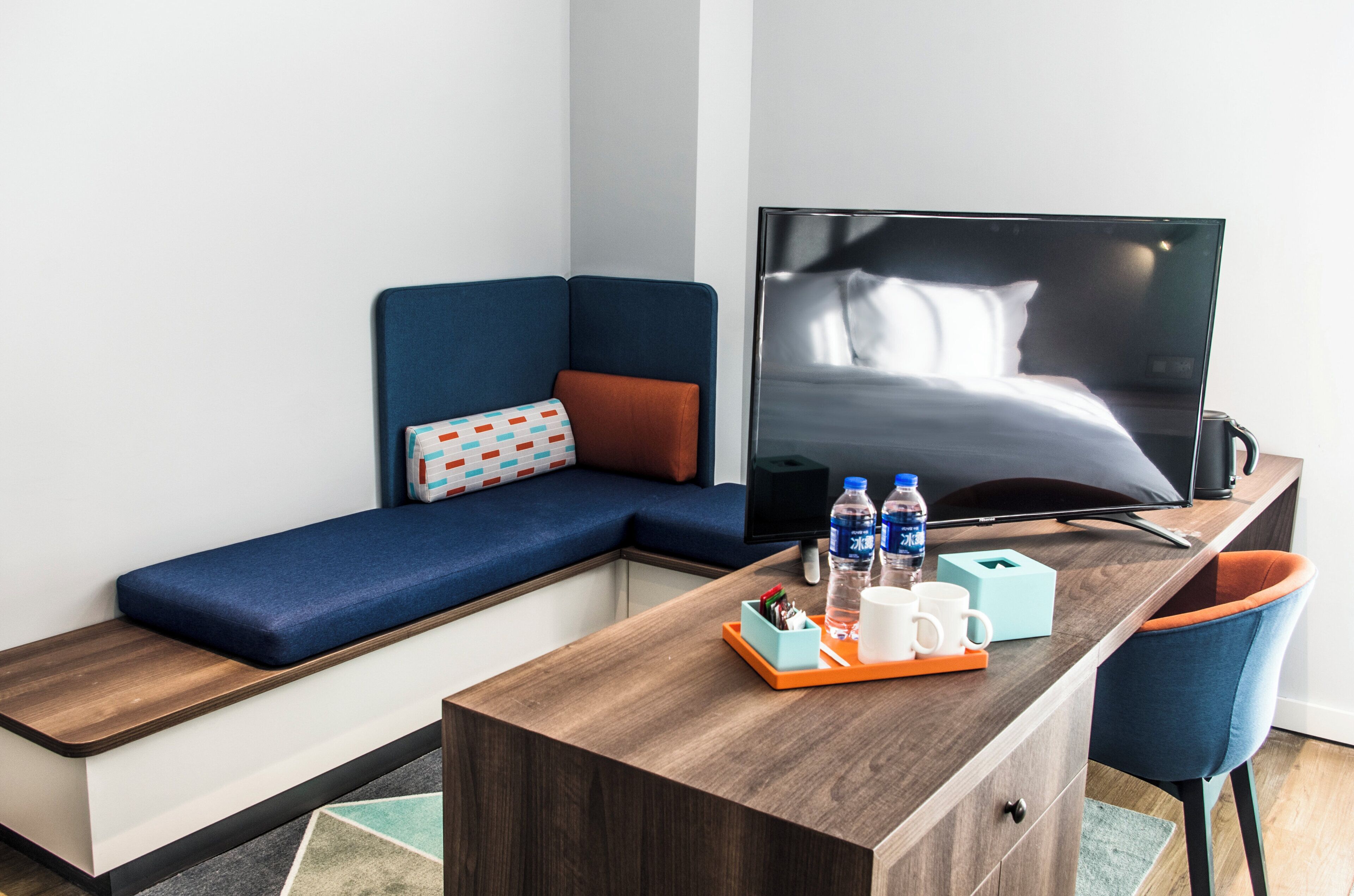 A modern hotel room with a sofa, a flat-screen TV, a wooden desk, and a chair.