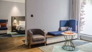 Una camera d'albergo moderna con un letto, una zona salotto con un divano e una poltrona blu, un tavolino con un vassoio di bottigliette d'acqua e una finestra vista fiori rosa.