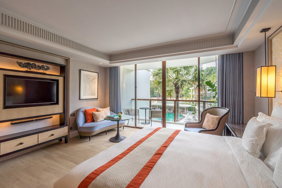 Une chambre d’hôtel avec un grand lit, une télévision, un canapé, un fauteuil, une petite table et une vue sur une piscine et un espace verdoyant.
