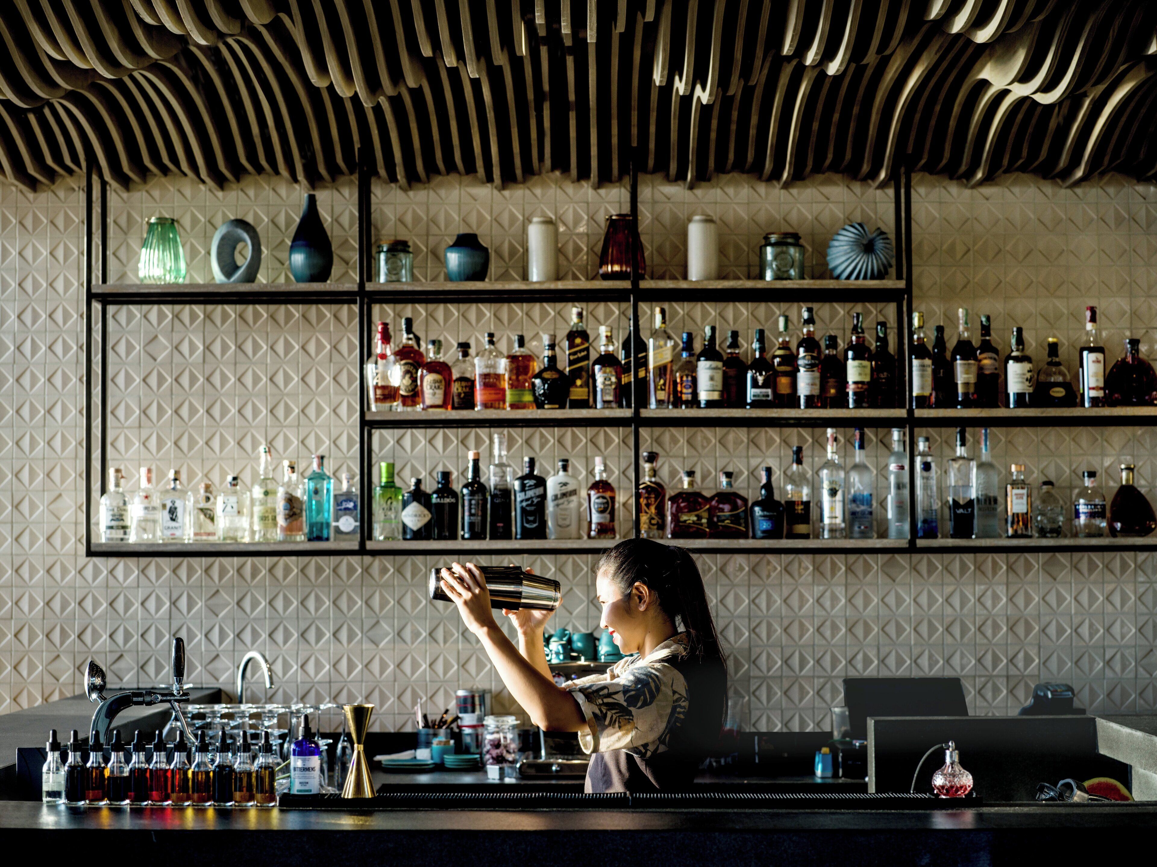A bar area with shelves of various liquor bottles, a bartender preparing a drink, and a unique ceiling design.