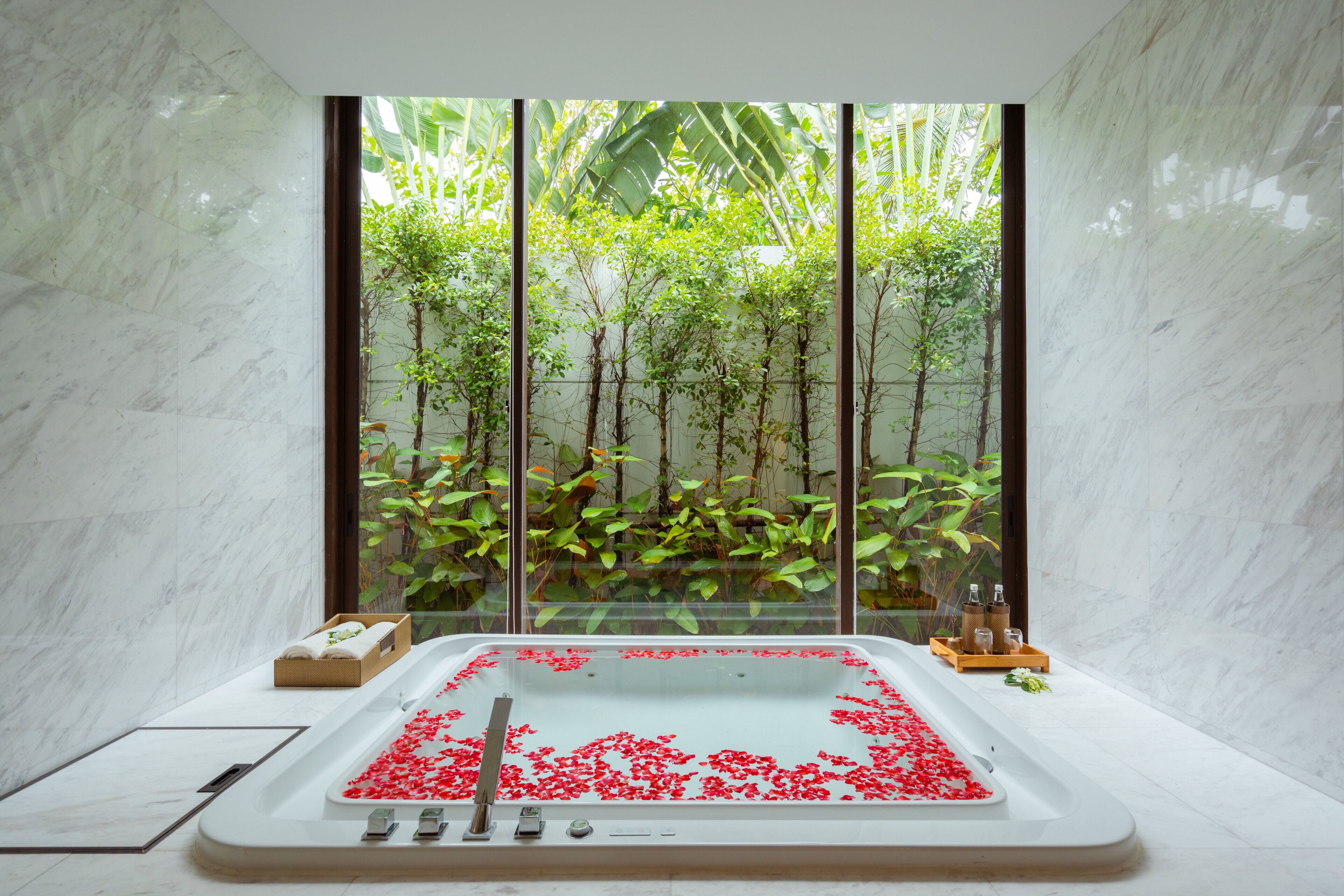 Une salle de spa dotée d’une grande baignoire remplie d’eau et de pétales de rose, entourée d’un mur de verre offrant une vue sur la verdure.