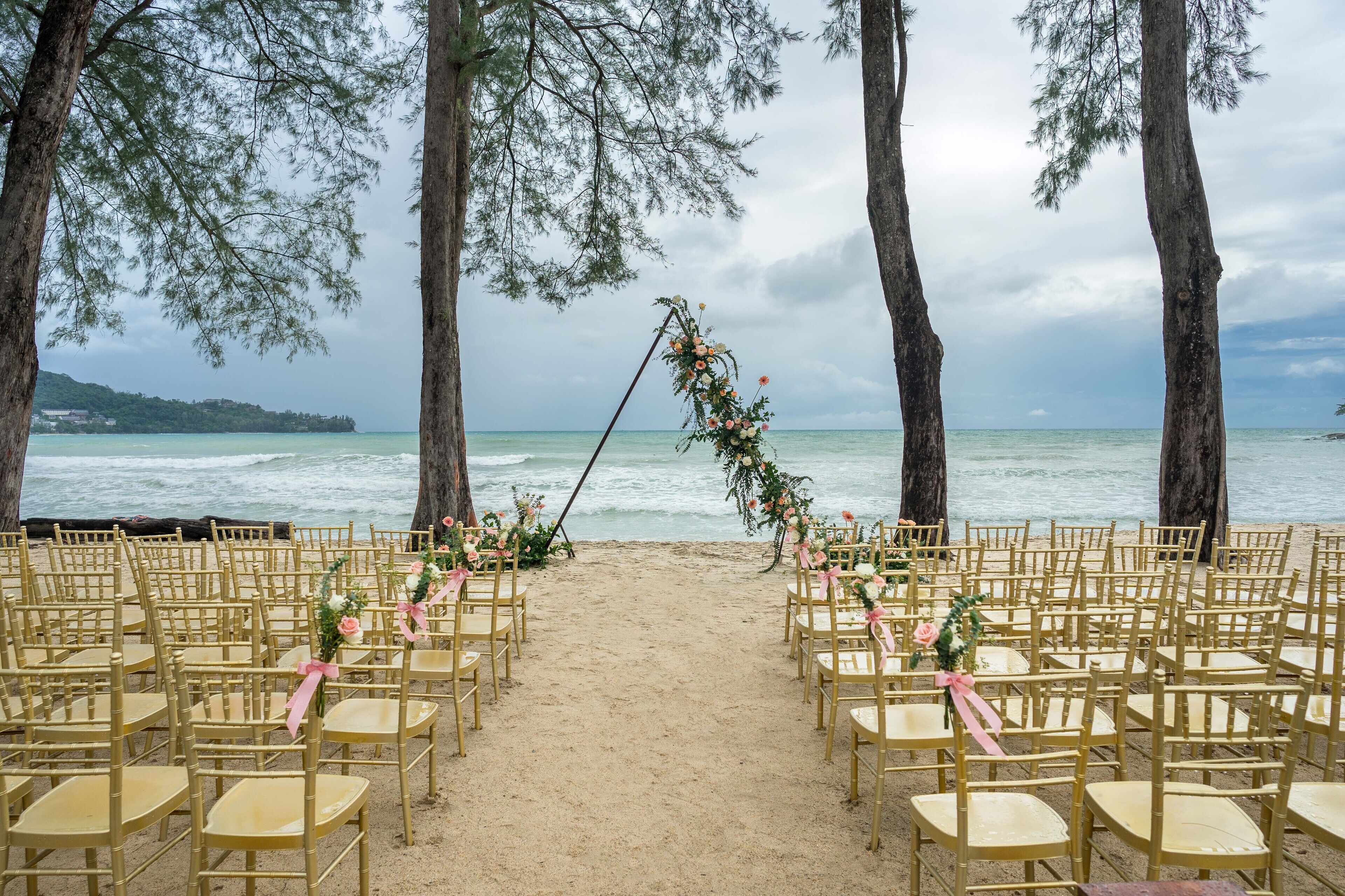 A beach wedding setup with rows of gold chairs and a floral arch.