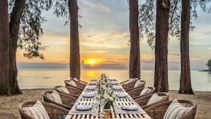 A long dining table set for two with wicker chairs, positioned on a beach with a sunset view.