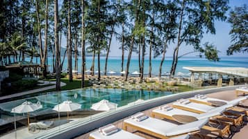 Area piscina di un resort con lettini e ombrelloni, circondata da alberi alti e con vista su una spiaggia.