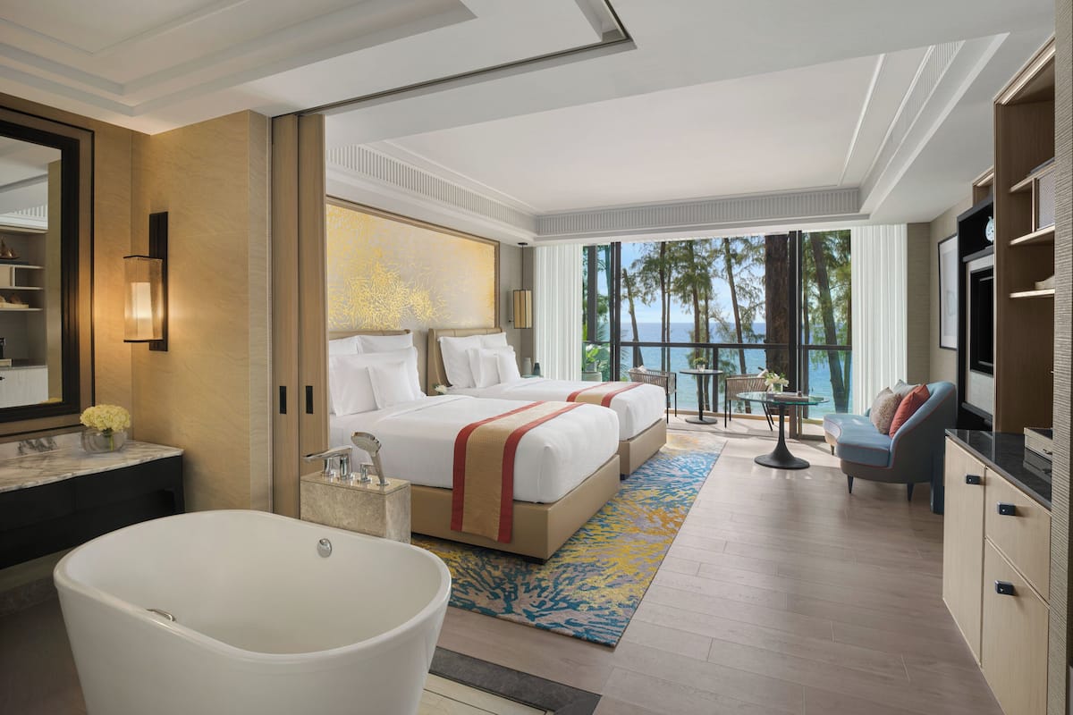 Een hotelkamer met een groot bed, een badkuip, een balkon met uitzicht op zee en een zithoek met een tafel en stoelen.