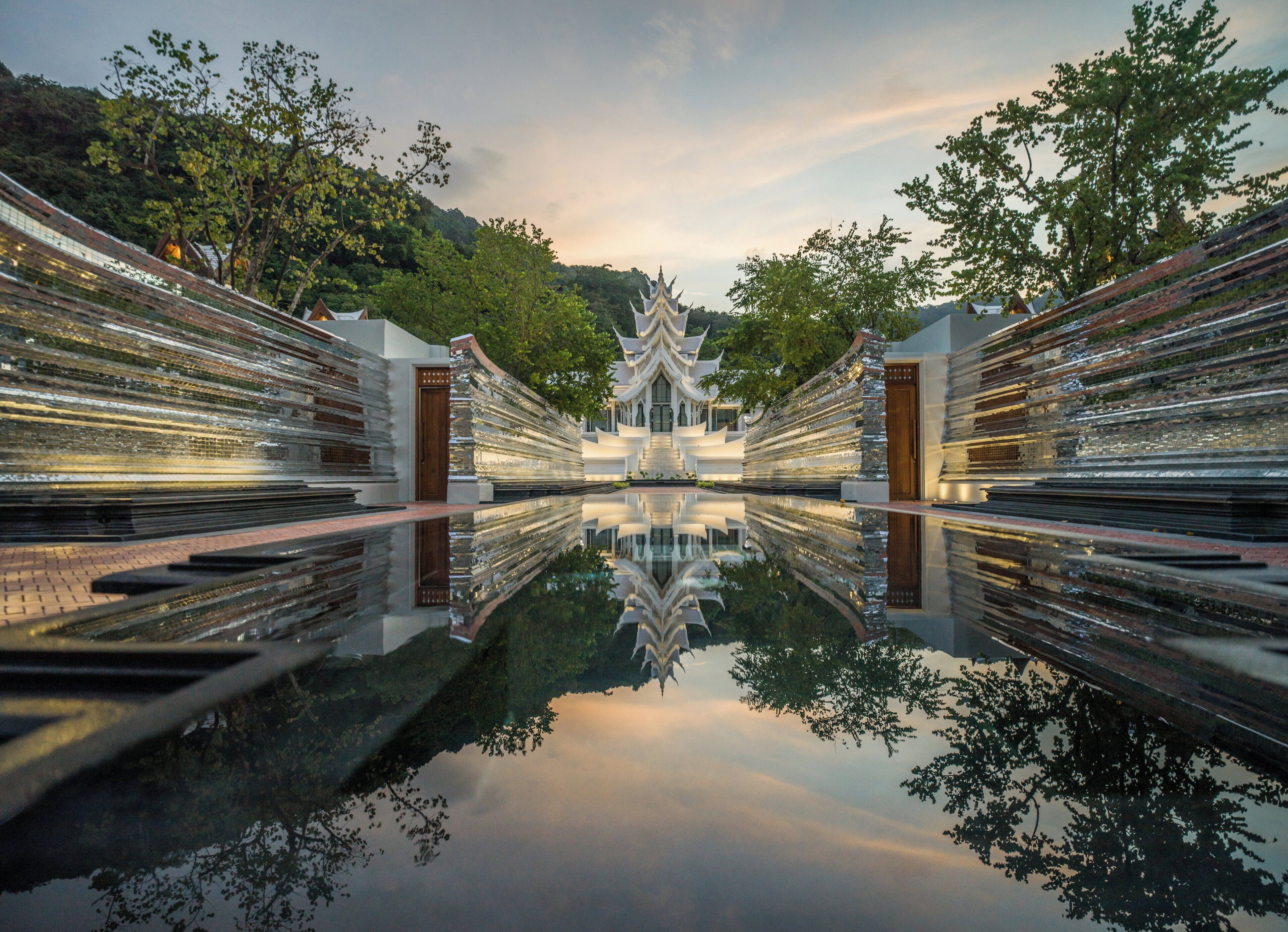 A symmetrical architectural structure with a central reflective water body, flanked by trees and a hilly backdrop.