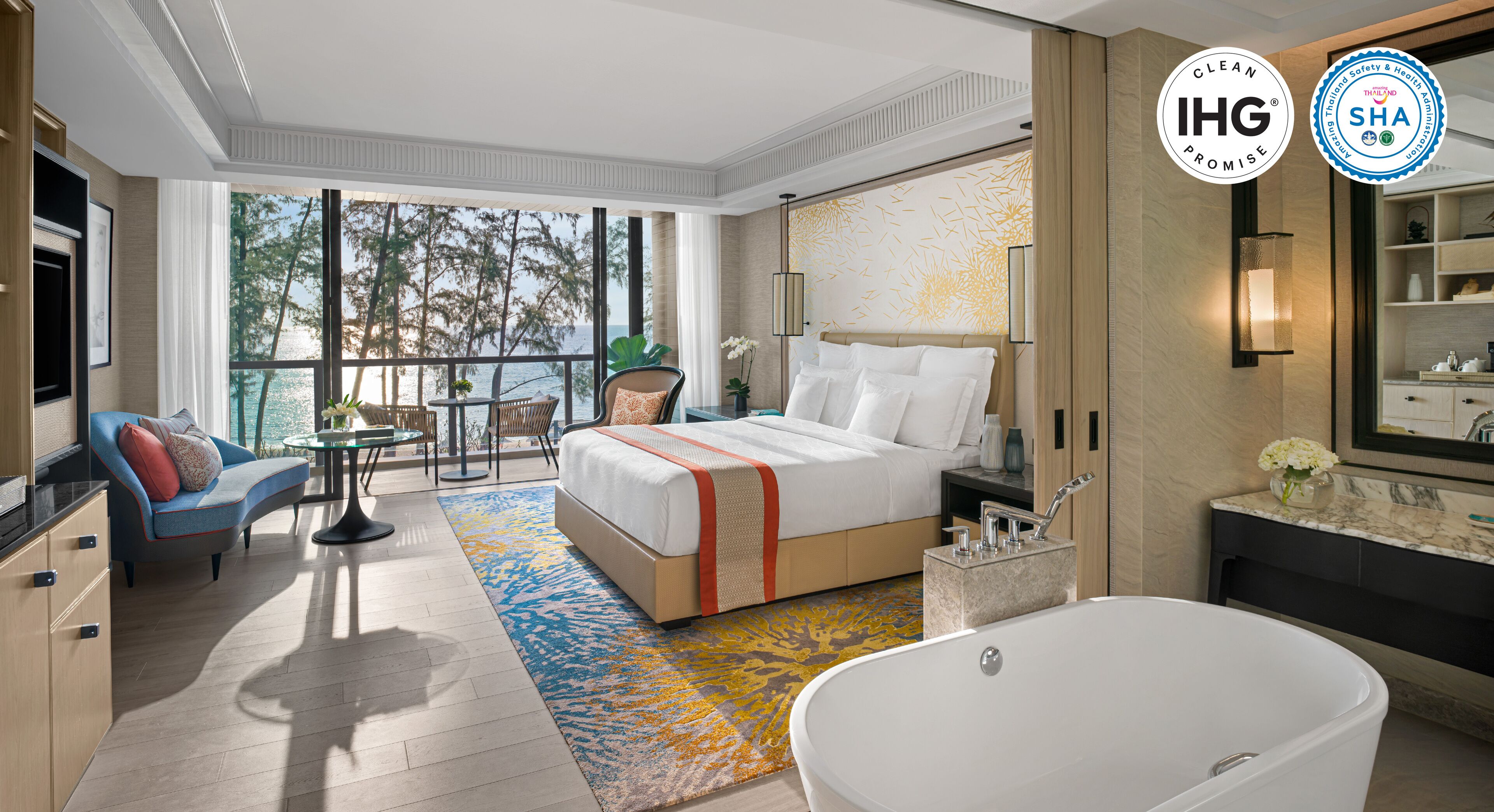 A modern hotel room with a large bed, a sitting area with a sofa and chairs, a dining table, a bathtub, and a view of the sea.