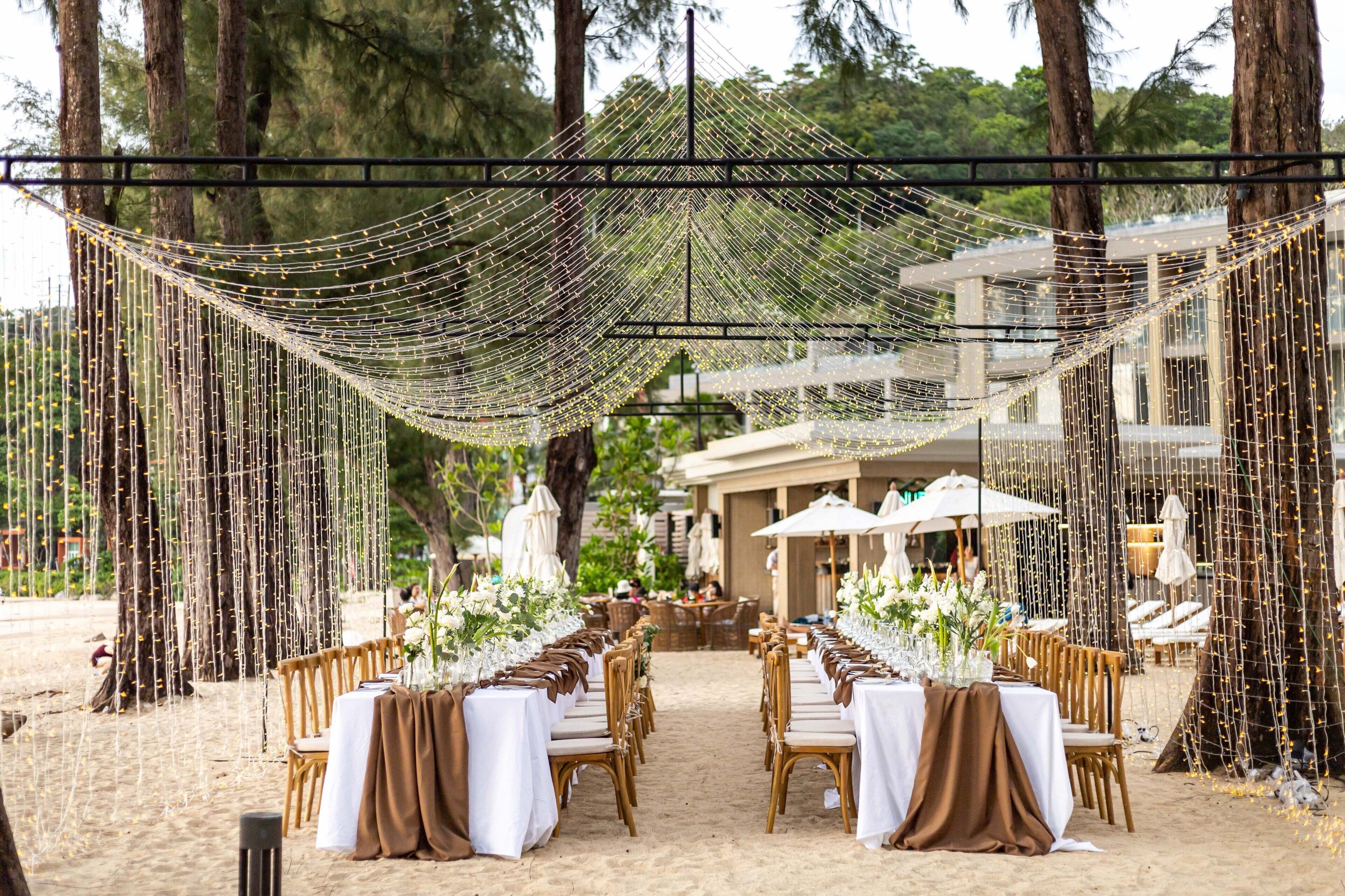 An outdoor dining area with tables set up on the sand, surrounded by trees and a building in the background.