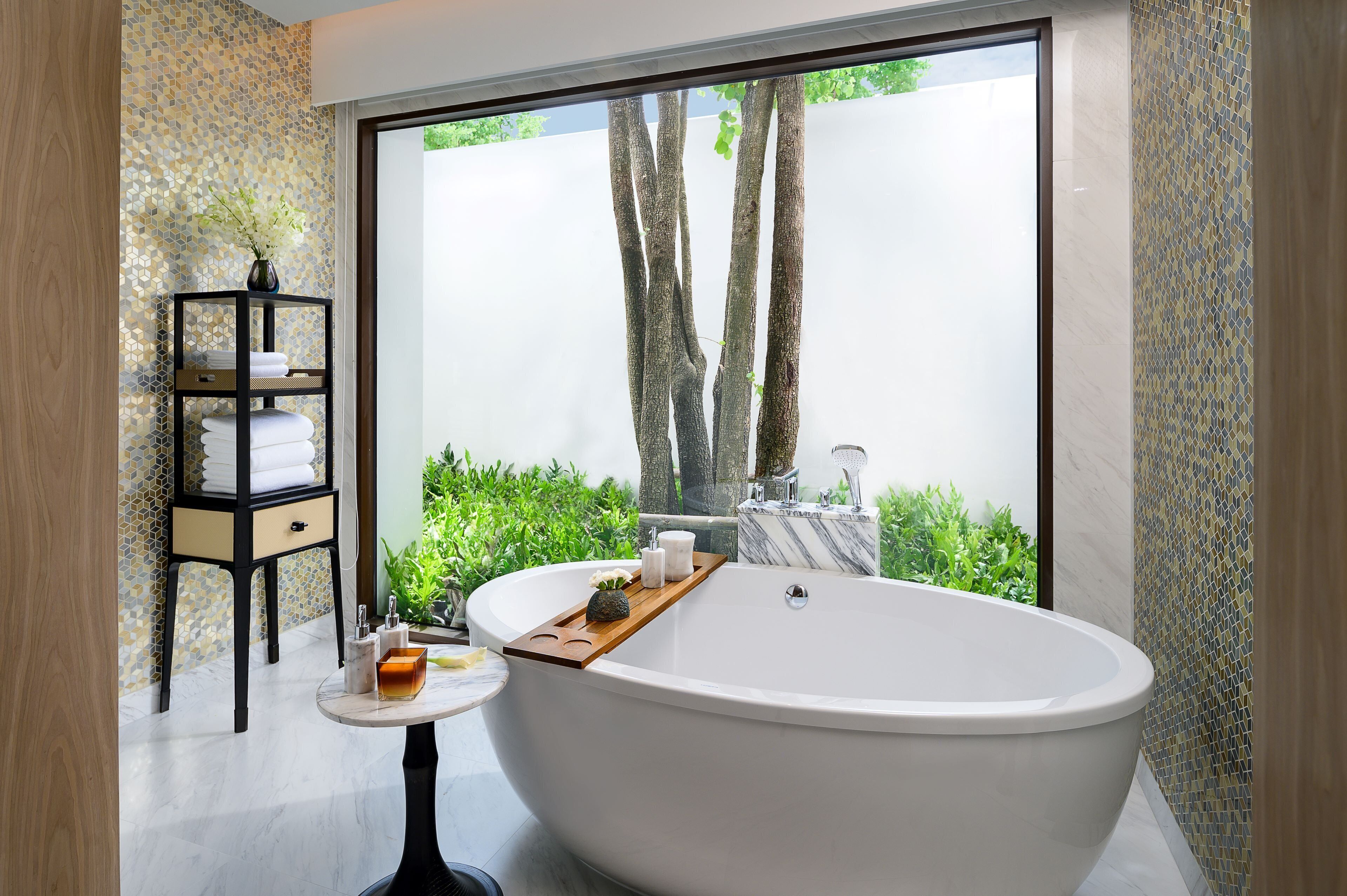 Een moderne badkamer met een vrijstaand bad, een klein tafeltje met toiletartikelen en uitzicht op groen buiten.