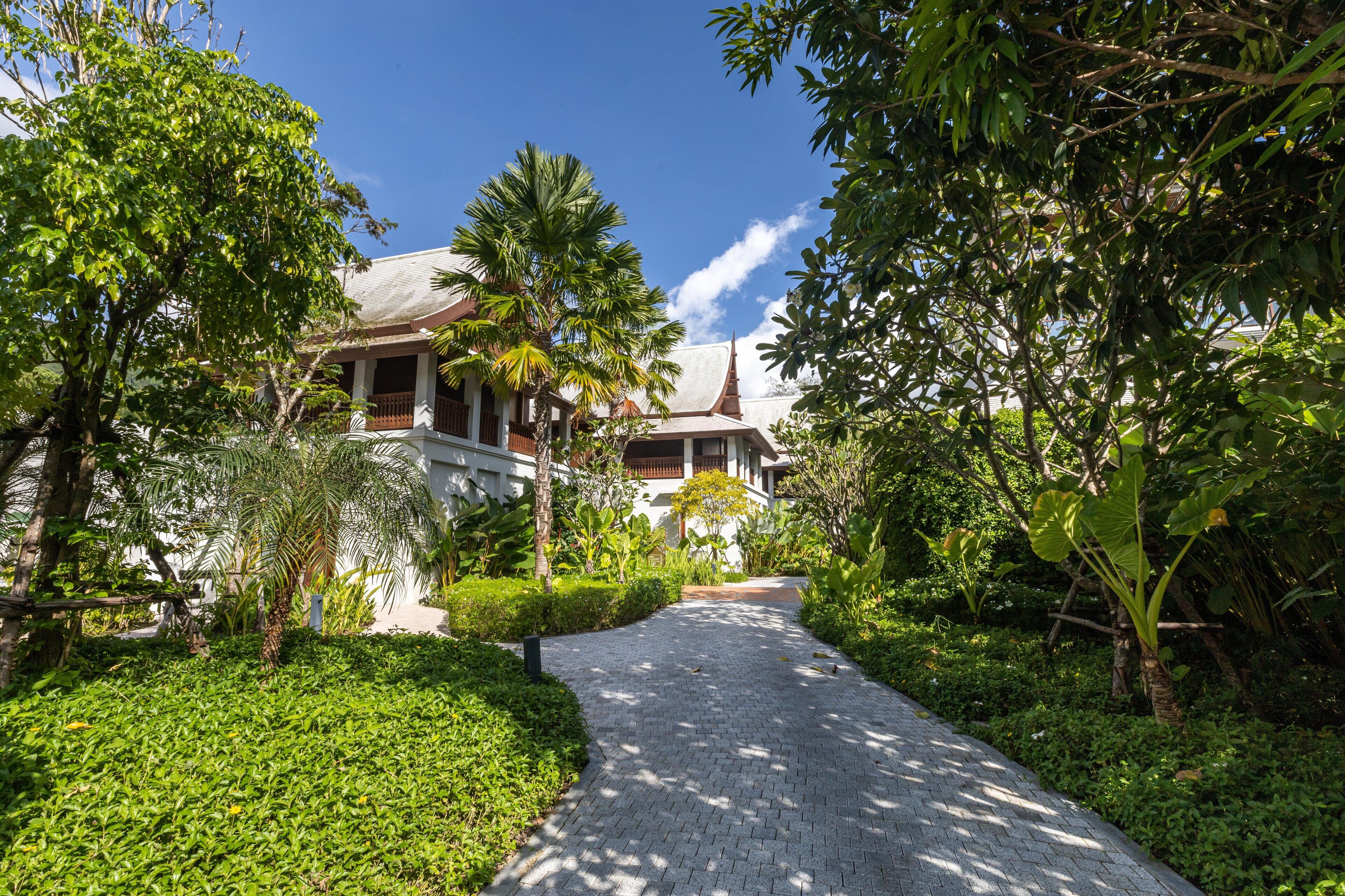 A pathway leading to a traditional building with a tiled roof and a spire, surrounded by lush greenery and palm trees.