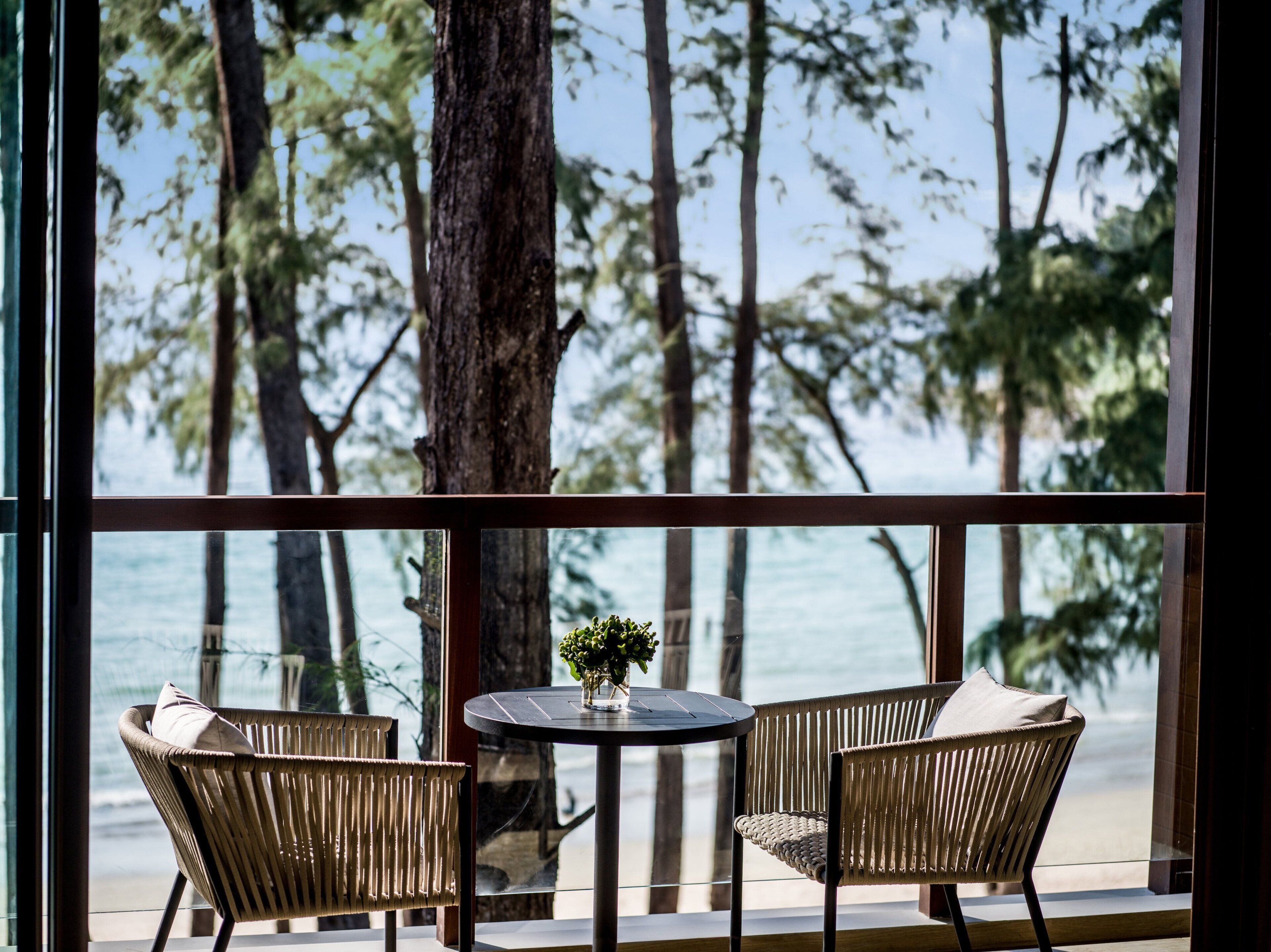 A balcony with two wicker chairs and a small table, overlooking a beach with trees and the ocean.
