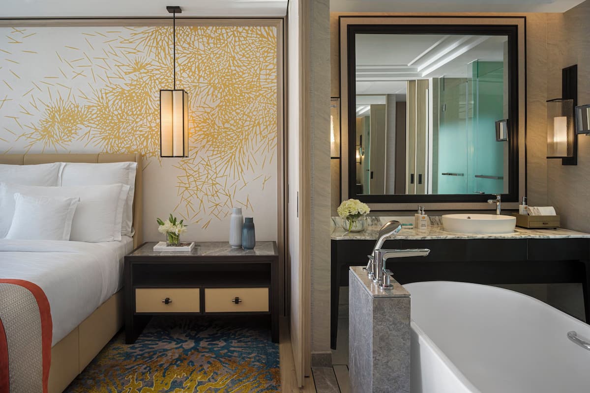 Een moderne hotelkamer met een groot bed, een nachtkastje met een bloevas, een badkamer met een grote spiegel, een badkuip en een marmeren aanrechtblad.