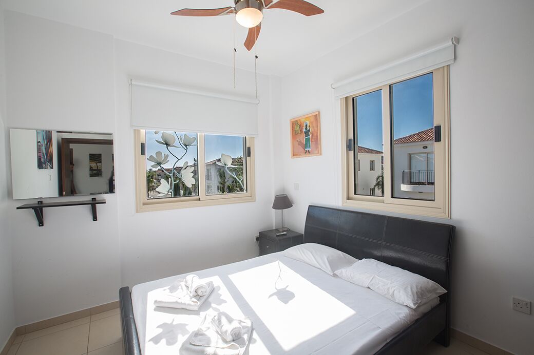 A bedroom with a bed, a ceiling fan, a window with a view, and a nightstand with a lamp.