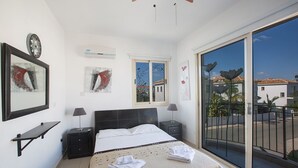 Une chambre à coucher comprenant un lit, des tables de chevet, un petit bureau et une grande fenêtre donnant sur des immeubles et des palmiers.