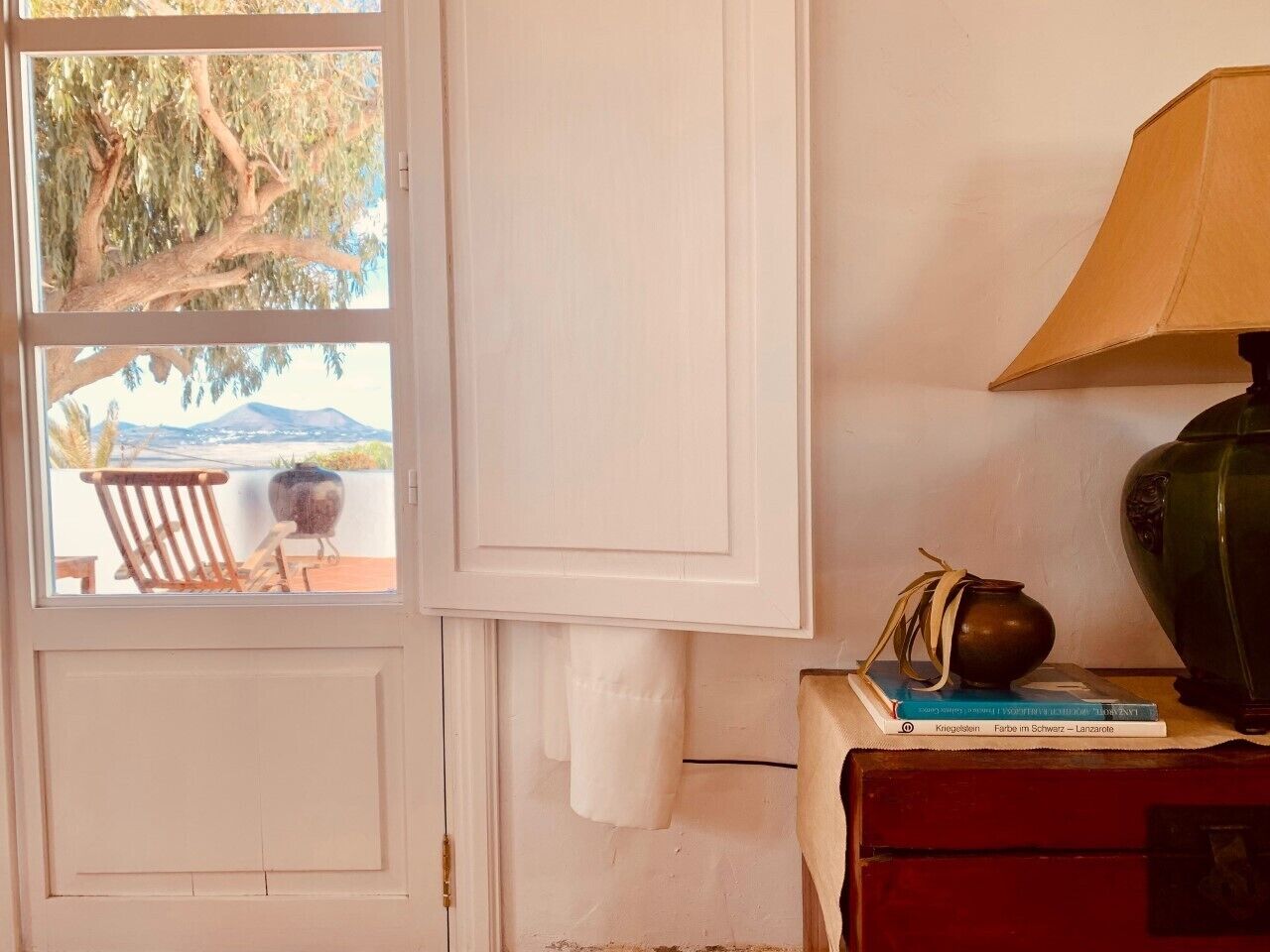 Una habitación con vista a una playa y montañas a través de una ventana, una lámpara sobre una mesa y un libro.