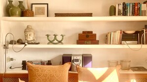 A bedroom with a bed, pillows, a bedside table with books and a radio, and a shelf with decorative items and books.