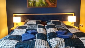 Ett sovrum med en mörkblå vägg, en stor säng med ett geometriskt mönstrat överkast, två sänglampor och en inramad tavla som föreställer en skog.