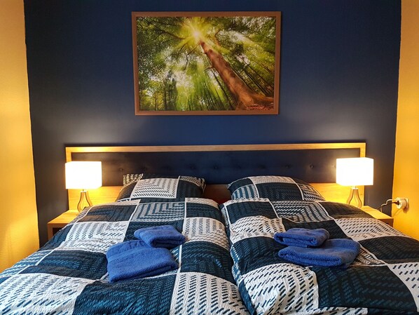 Un dormitorio con una pared azul oscuro, una cama grande con edredón de estampado geométrico, dos lámparas de noche y un cuadro enmarcado de un bosque.