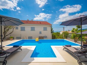 A rectangular swimming pool with a clear blue water, surrounded by lounge chairs and umbrellas, with a building in the background.