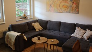 A modern living room with a dark grey sectional sofa, two round wooden side tables, a beige throw blanket, and a colorful abstract painting on the wall. - Apartment Annette Mieth - Apartment (Weitenhagen)