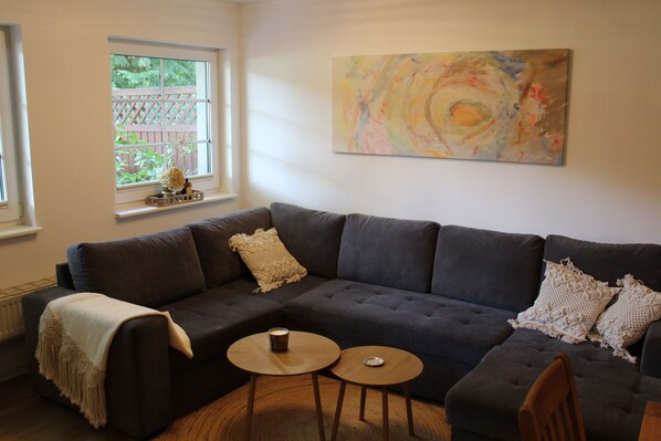 A modern living room with a dark grey sectional sofa, two round wooden side tables, a beige throw blanket, and a colorful abstract painting on the wall.