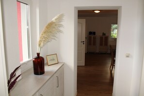 A hallway with a wooden floor, a white wall with a pink stripe, a vase with pampas grass, and a framed picture on a cabinet.