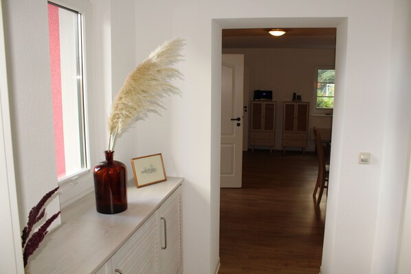 A hallway with a wooden floor, a white wall with a pink stripe, a vase with pampas grass, and a framed picture on a cabinet.