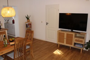 A living room with a wooden dining table, chairs, a television on a wooden cabinet, a bookshelf, and a hanging lamp.