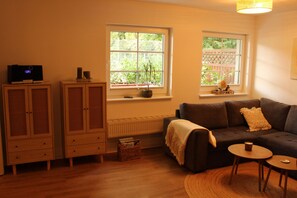 Ein gemütliches Wohnzimmer mit einer Couch, einem kleinen Tisch und einem Schrank. Es gibt zwei Fenster mit Pflanzen auf den Fensterbänken.