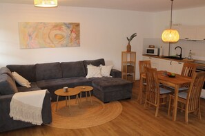 A modern living room with a grey sectional sofa, a small wooden dining table with four chairs, and a kitchen area with white cabinets and a microwave.