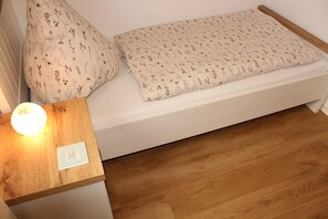 Ein ordentlich gemachtes Bett mit blumigem Bettzeug und Kissen, ein Nachttisch aus Holz mit einer Lampe und eine weiße Wand.