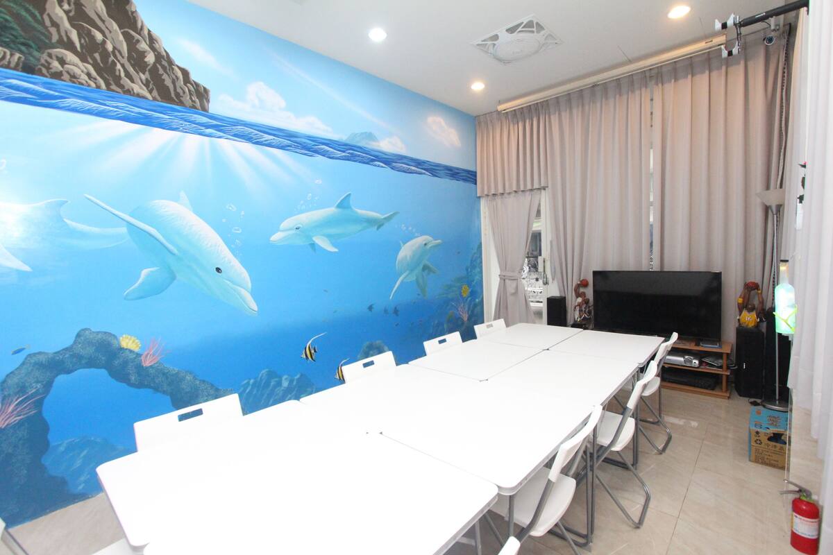 A room with a large mural of dolphins and an underwater scene on the wall, a white table with chairs, a flat-screen TV, and a fire extinguisher on the floor.