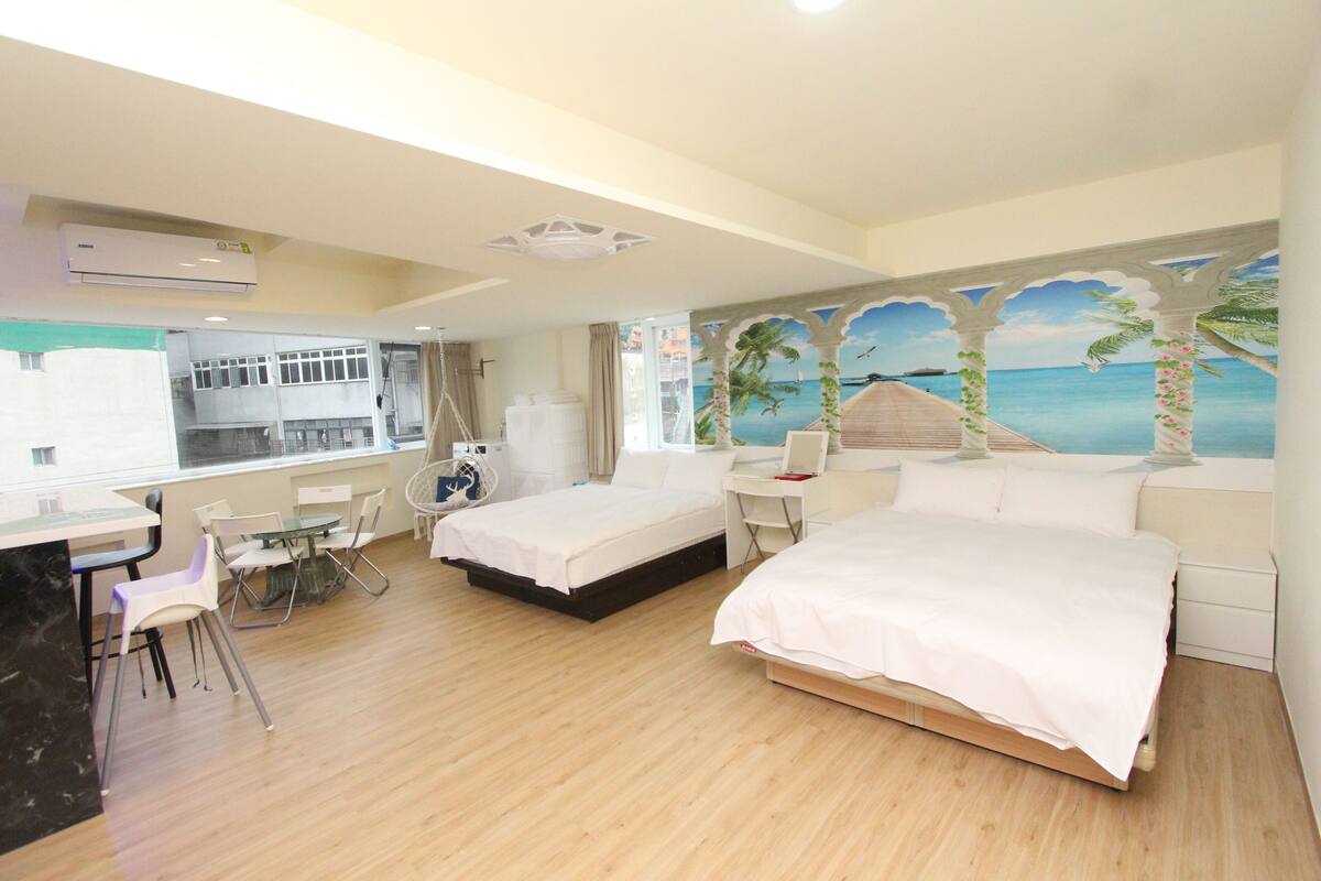 A hotel room with two beds, a desk, a chair, a dining table, and a mural of a beach scene.