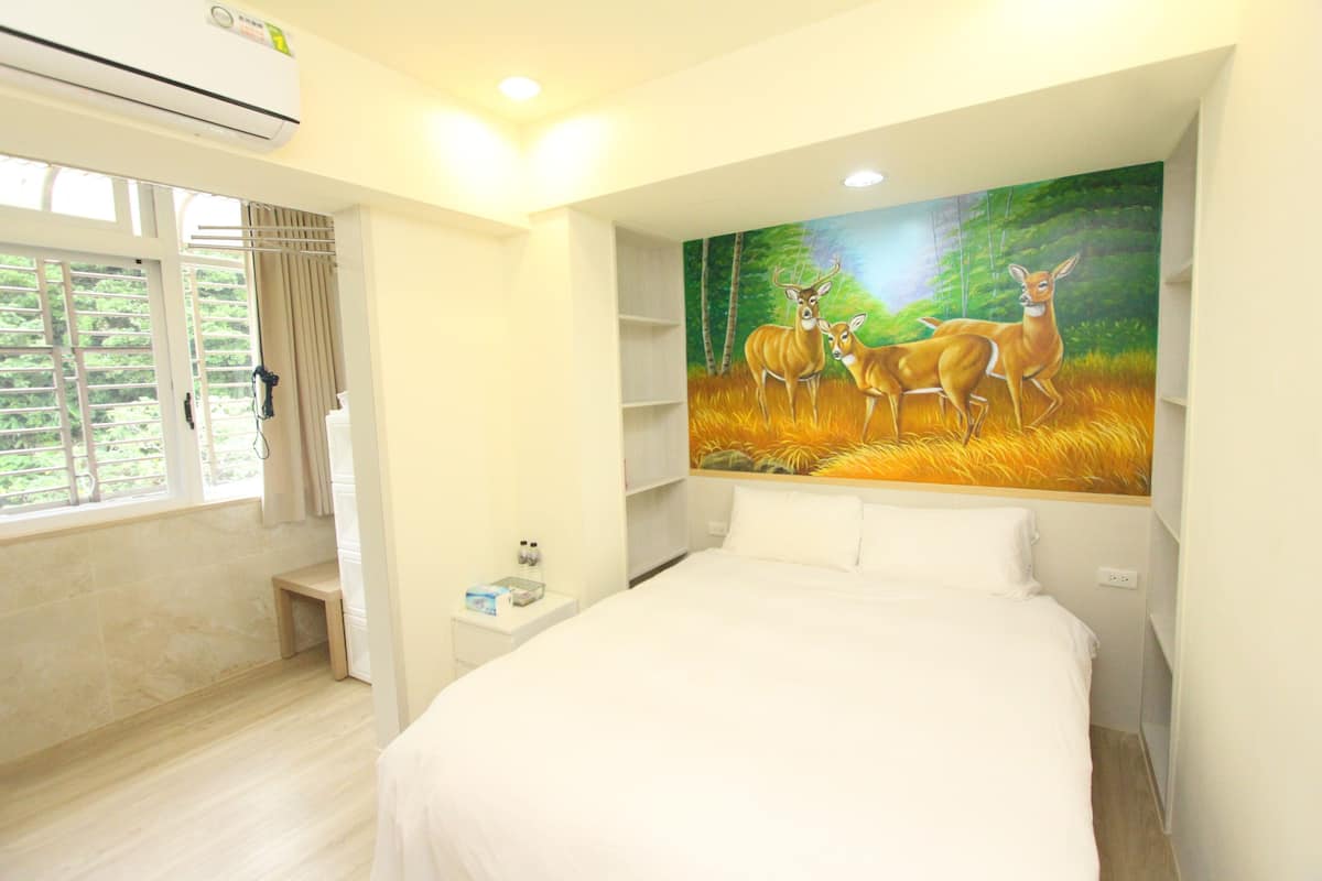 A bedroom with a large bed, a window with curtains, a painting of deer, and a small bedside table.