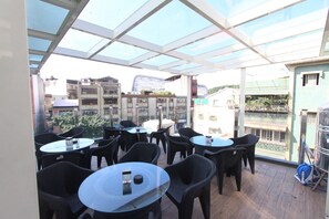 An outdoor patio area with round tables and chairs, a glass ceiling, and a view of buildings in the background. - HF Keelung (Keelung)