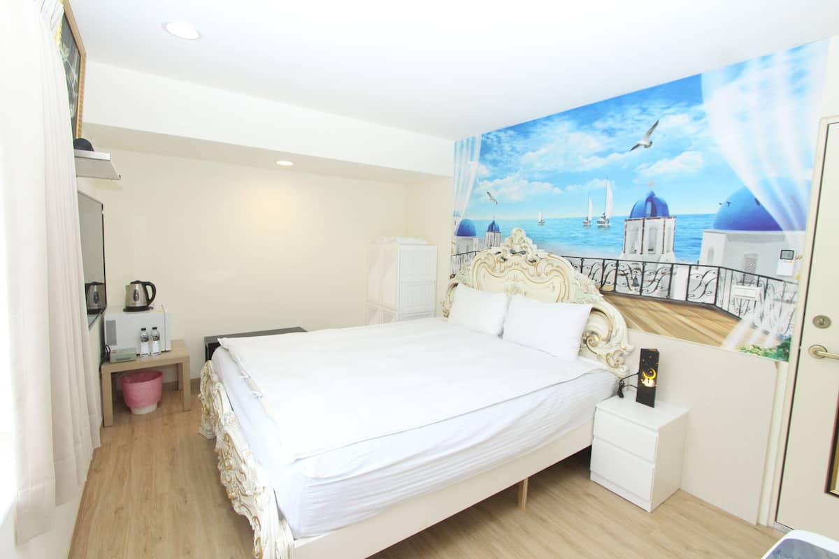 A bedroom with a large bed, a nightstand, a wall-mounted TV, and a painting of a coastal scene.