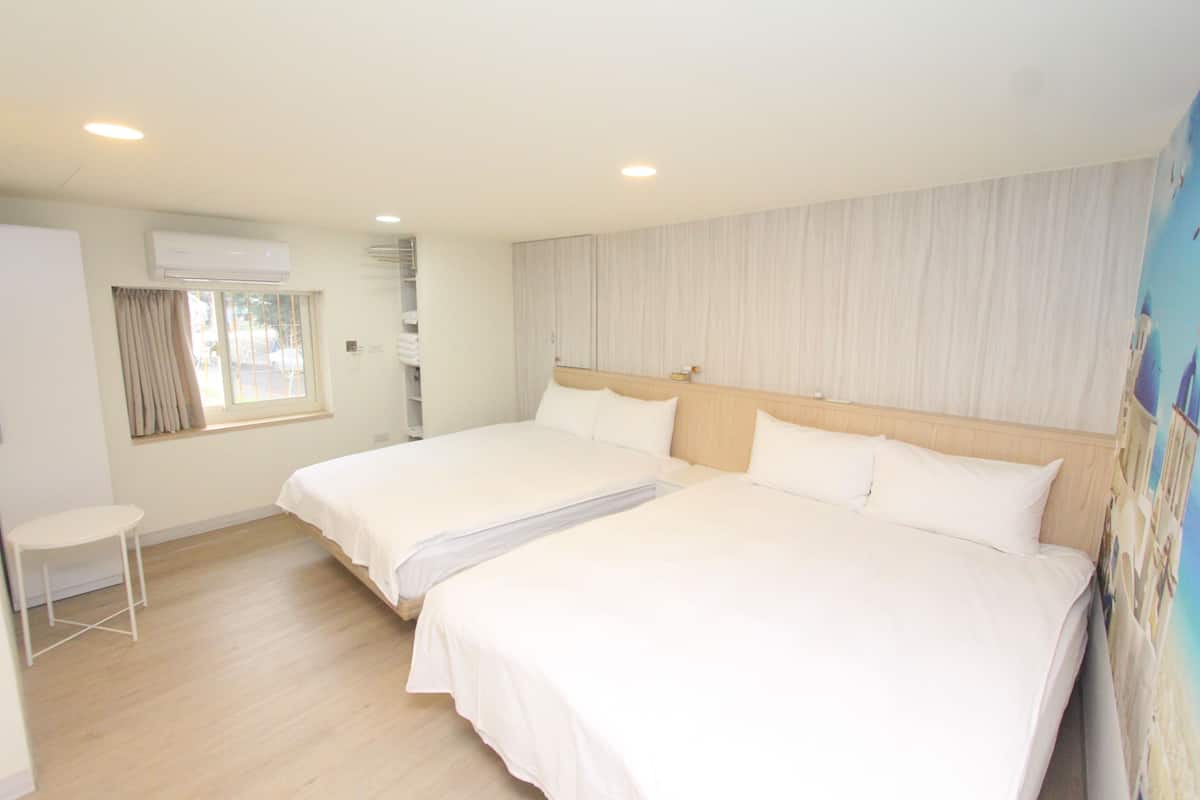 A hotel room with two beds, a small table, an air conditioning unit, and a window with curtains.