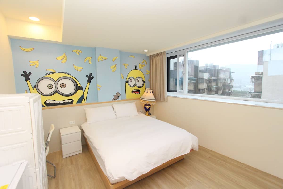 A hotel room with a bed, a nightstand, a lamp, and a window with a city view.
