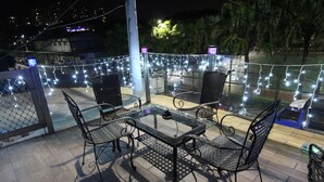 Een terras buiten met een tafel met glazen blad en vier stoelen, omringd door lichtslingers en een uitzicht op een stadsbeeld bij nacht.