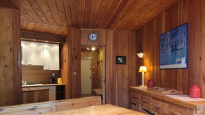 A wooden interior with a dining table, benches, a kitchen area, and a clock on the wall.