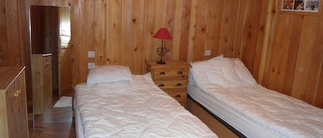 A wooden-paneled room with two beds, a nightstand with a lamp, and a dresser.