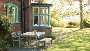 Una casa de ladrillo con un ventanal verde, una mesa con sillas y un banco en el exterior.