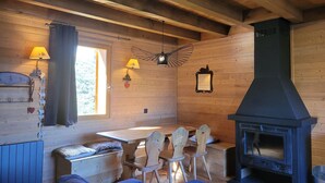A cozy cabin interior with a dining area, wooden walls, a fireplace, and a window with curtains.
