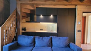 A modern living room with a blue sofa, a wooden coffee table, and a kitchen area with dark cabinets and a built-in oven.