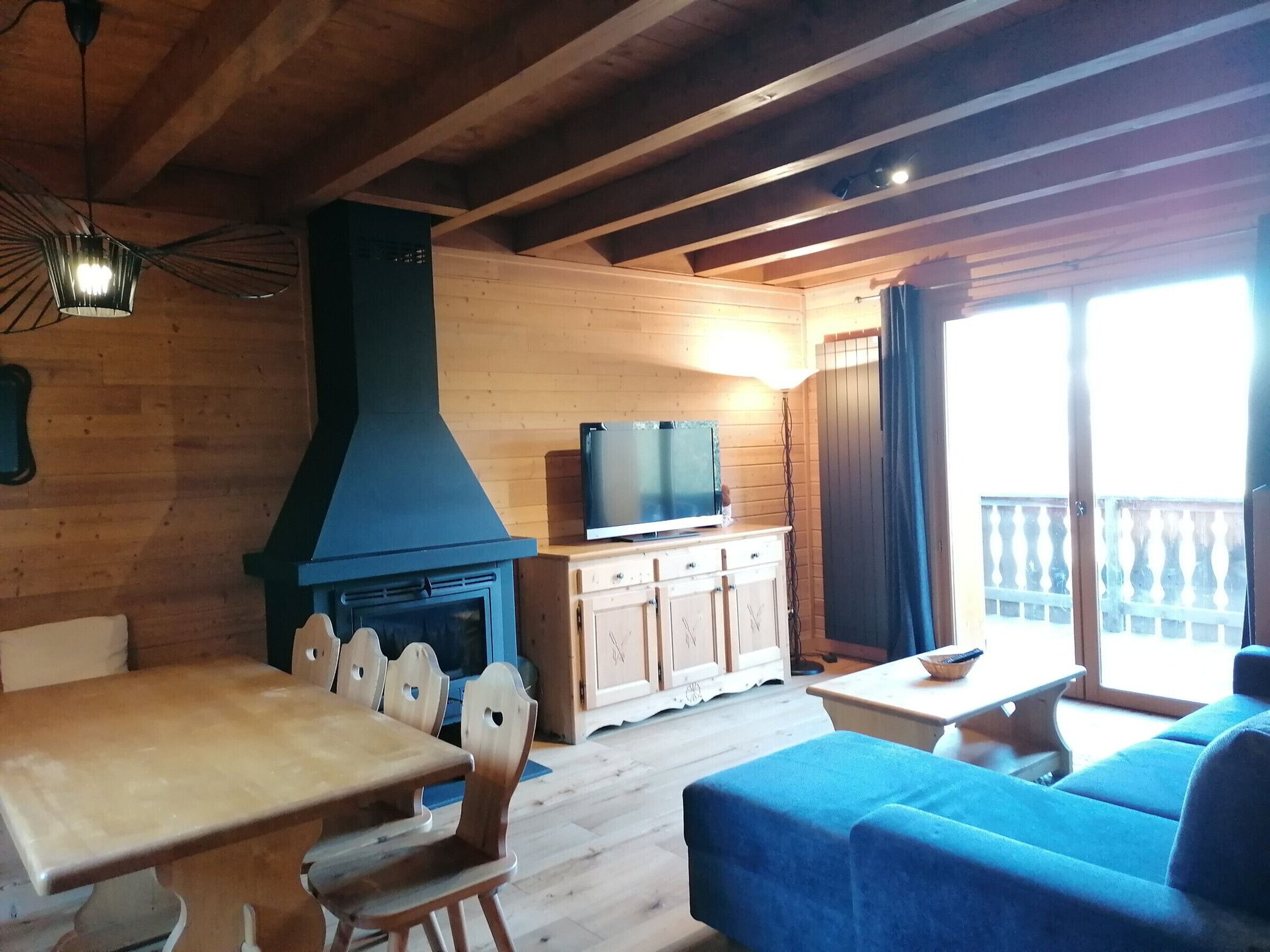 Een houten interieur met een eettafel, stoelen, een bank, een televisie op een kast, en een balkon met een hekwerk.