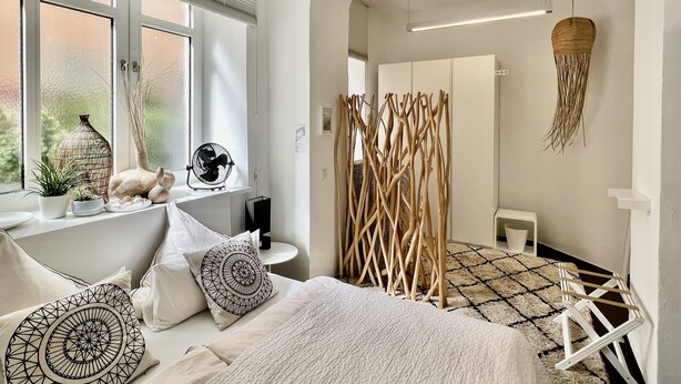 A modern bedroom with a large bed, a bedside table with decorative items, a window with blinds, and a view of the outside. - Best deal in town !  Cocoma Designer Apt deluxe in the center of Munich (Ludwigsvorstadt-Isarvorstadt)
