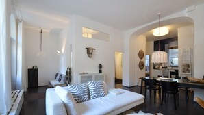 A modern living room with a white sofa, a dining area with a table and chairs, and a kitchen in the background. - Best deal in town !  Cocoma Designer Apt deluxe in the center of Munich (Ludwigsvorstadt-Isarvorstadt)